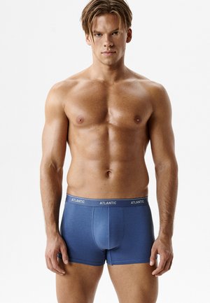 Muskularny mężczyzna stojący twarzą do przodu, ubrany w niebieskie bokserki marki Atlantic na jednolitym białym tle.