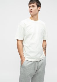 Bijela pamučna majica s kratkim rukavima, okruglim izrezom, s diskretnim logotipom na prsima. Uparen s svijetlosivim trenirkama.