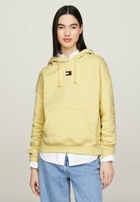 Felpa gialla con tasca a marsupio, cordini e una patch con logo. Indossata sopra una camicia bianca con colletto e abbinata a jeans chiari.