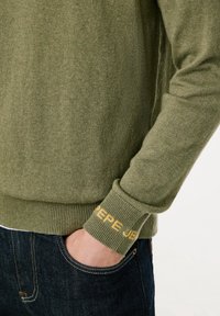 Personne portant un pull vert olive avec le texte « PEPE JEANS » sur le poignet, main dans la poche d'un jean bleu foncé.