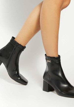 Black leather ankle boots with a square toe, side zipper, low block heel, and gold chain accent on the cuff. Smooth texture and sleek design.