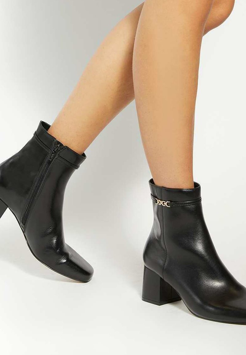 Black leather ankle boots with a square toe, side zipper, low block heel, and gold chain accent on the cuff. Smooth texture and sleek design.