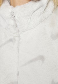 Bílá falešná kožešinová bunda s měkkým, texturovaným povrchem, s předním zipem a vysokým límcem. Výrazná lehkost designu.