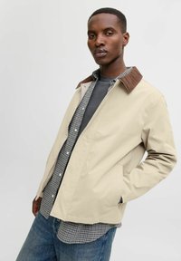 Beige jakke med en brun kordfløjlskrage, lange ærmer og sidelommer, lagd over en ternet skjorte. Jeansbukser er synlige.