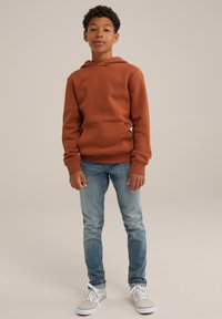 Felpa color ruggine con tasca a marsupio, abbinata a jeans azzurri chiari e scarpe da ginnastica grigie. Il tessuto sembra morbido e ha una vestibilità comoda.
