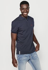 Tamsiai mėlynas trumpomis rankovėmis polo marškinėliai su tekstūriniu raštu, dviem mygtukais ir kontrastingais apvadais ant apykaklės ir rankovių.