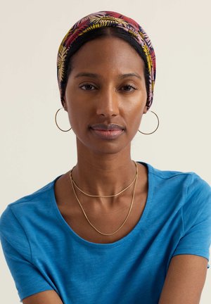 Sötét bőrű nő, kék inget visel, arany karika fülbevalókat, réteges arany nyakláncokat és színes mintás fejkendőt, előre néz.