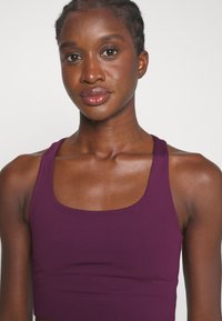 Violeta sporta kūrts ar platām siksnām un kvadrātveida kakla izgriezumu, izgatavots no gludas auduma, ar pieguļošu siluetu un smalkām šuvēm.