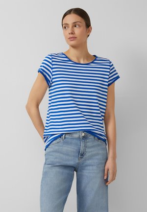 Frau trägt ein blau-weiß gestreiftes kurzärmeliges Shirt und hellblaue Jeans, steht mit einer Hand hinter dem Rücken.
