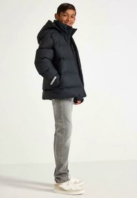 Zwarte pufferjas met capuchon, voorzien van een gewatteerd ontwerp en grijze accenten, gecombineerd met lichtgrijze spijkerbroek en witte sneakers.