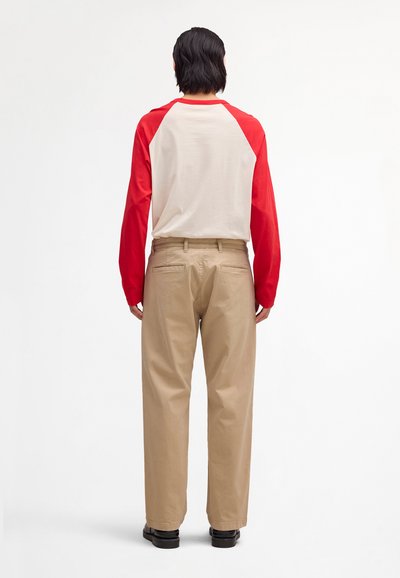 Άτομο που φαίνεται από πίσω, φορώντας μπεζ παντελόνια, κόκκινο και λευκό πουκάμισο με μακριά μανίκια και μαύρα παπούτσια, μπροστά σε λευκό φόντο.