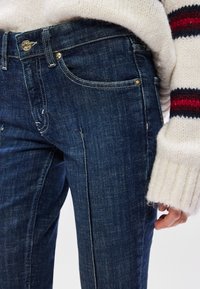 Jeans de ganga azul escuro com um design clássico de cinco bolsos, hardware dourado e uma textura subtil. Combinados com um suéter de malha creme.