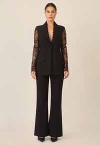 Schwarzer Anzug mit langärmeliger Spitzenoverlay und einer taillierten Blazerjacke mit tiefem V-Ausschnitt, kombiniert mit ausgestellten Hosen und High Heels.