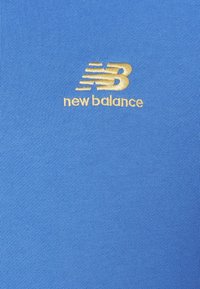 Sininen urheilupaita, joka on valmistettu sileästä kankaasta ja jossa on kullanvärinen kirjailtu "New Balance" -logo ylävasemmalla puolella.