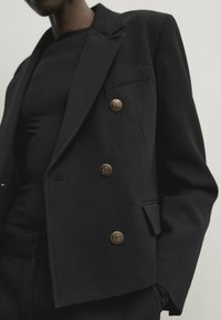 Musta kaksirivinen bleiseri kultaisilla napin koristeilla, lovellut kaulukset ja teksturoitu kangas; sivutaskut ja räätälöity istuvuus.