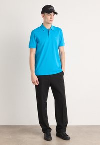 Lyseblå polo-skjorte med krage, kombinert med svarte bukser og svarte sneakers. Skjorten har et logo på brystet og korte ermer.