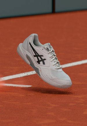 Valge spordijalats musta logoga, mis tõstab punast savipuru tenniseväljaku lähedal valgete piirjoonte kõrval.