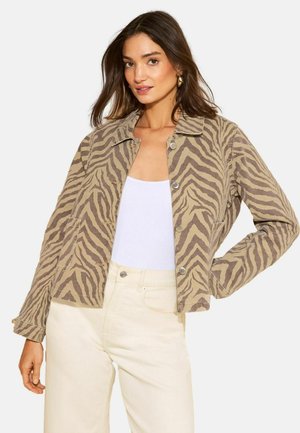 Naine kannab beeži ja pruuni sebratriibulist jopet valge pluusi ja kreemjate kõrge vöökohaga pükstega, poseerides ühe käega puusal.