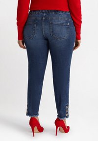 Mörkblå denimjeans med elastisk midja, med fransade fållar och gyllene knappaccenter, bärs tillsammans med röda högklackade skor.