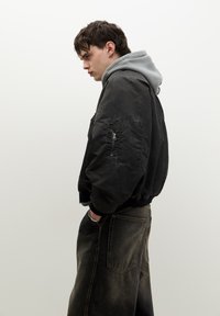 Čierna bomber bunda s kapucňou prehodená cez šedú mikinu, so zipsovým vreckom na prednej strane a rebrovanými manžetami, v kombinácii s tmavými denimovými nohavicami.