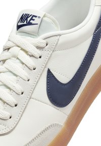 Sapatilha de pele branca com um swoosh azul-marinho, língua texturizada e atacadores em creme. A sola é de borracha com uma tonalidade bege.