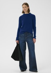 Modrý pletený sveter so zdobeným výstrihom, skombinovaný s vysokými džínsami širokých nohavíc a čiernou taškou typu tote.