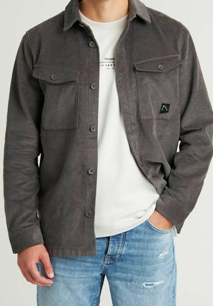 Hombre con camisa abotonada de pana gris oscuro sobre camiseta gráfica blanca y vaqueros azul claro desgastados, mano en el bolsillo.