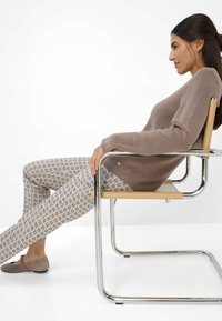 Pull en tricot marron associé à un pantalon clair à motifs présentant des designs géométriques. Modèle assis sur une chaise en métal minimaliste.