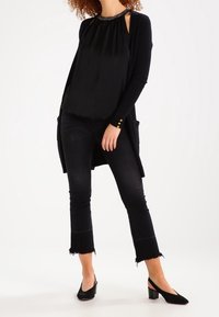 Svart, ärmlös topp med rynkad halsringning; lager på lager med en lång svart cardigan; matchad med korta svarta jeans; svarta pumps med spetsig tå.