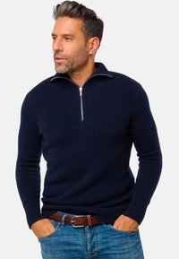 Pull-over en tricot bleu marine avec une fermeture éclair quart, des poignets côtelés et un motif texturé sur la poitrine. Porté sur un jean en denim bleu clair.