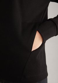 Main insérée dans la poche latérale d'un vêtement noir à manches longues montrant la texture du tissu et le détail de la poche.