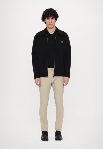 Sort lynlåsjakke med krave opdelt over en sort polo, parret med beige slim-fit bukser og sorte slip-on sko.