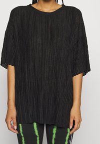 Top oversized plissettato nero con scollatura rotonda e maniche corte, caratterizzato da linee verticali per texture. Abbinato a pantaloni verde tie-dye.