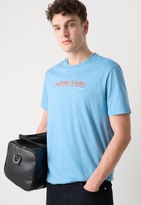 Šviesiai mėlynas medvilninis marškinėliai su apvalia iškirpte ir raudonu "Calvin Klein" logotipu atspausdintu per krūtinę. Juoda odinė rankinė laikoma kairėje rankoje.
