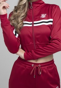 Bordeaux polyester træningsdragt med en lynlåslukket jakke med hvide og sorte striber samt matchende bukser med elastisk talje og snøre.