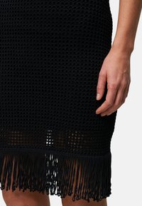 Robe tricotée noire avec un motif lâche et texturé. Présente un détail d'ourlet frangé et une silhouette ajustée. Main visible sur le côté.
