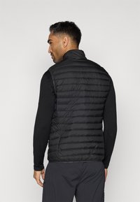 Μαύρη επενδυμένη γιλέκα με ψηλό γιακά, στενό καπιτονέ σχέδιο και λεία υφή, φορεμένη με μια μαύρη μπλούζα μακρυμάνικη από κάτω.