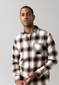 Camicia flannel con bottoni in crema, nero e marrone a quadri. Caratterizzata da una tasca sul petto, colletto classico e tessuto morbido e testurizzato.