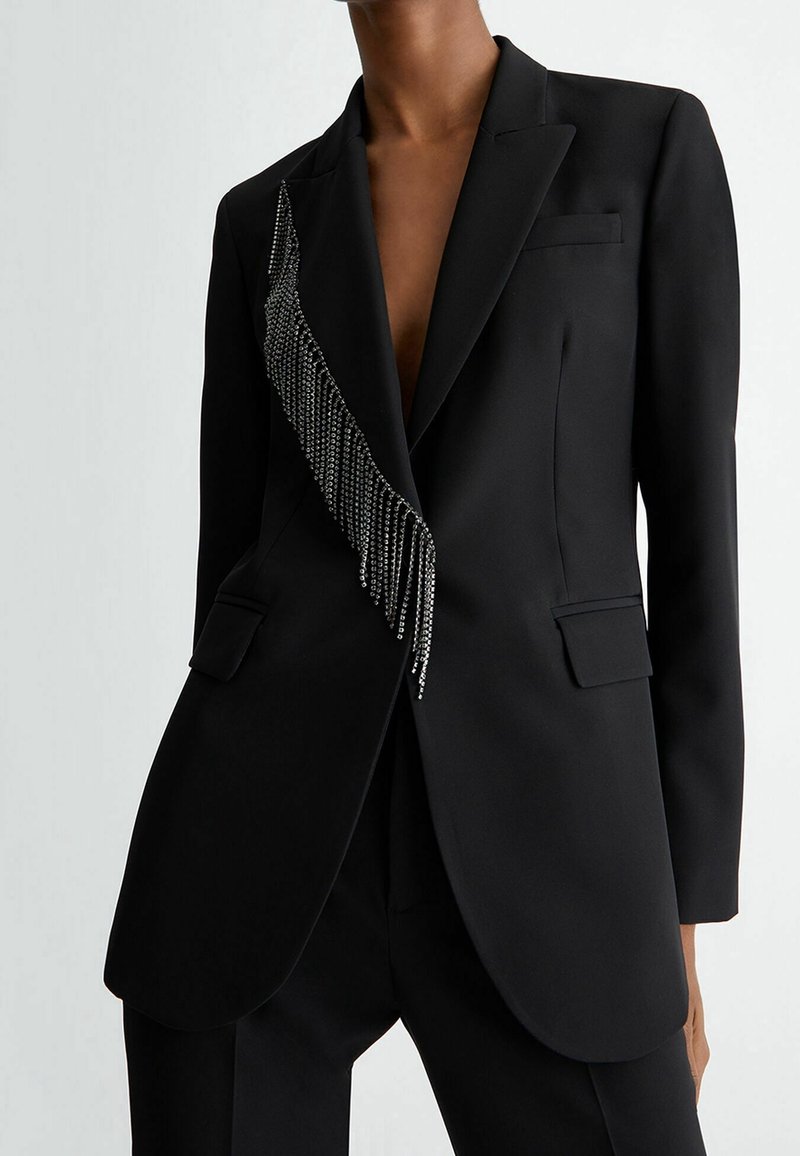 Blazer noir cintré avec des franges argentées le long d'un revers, porté ouvert sur un pantalon noir assorti sur une personne debout.