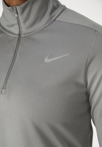 Szürke sportfelső, sima, rugalmas anyagból készült. Magas gallérral, félcipzárral és egy diszkrét, fényvisszaverő Nike logóval rendelkezik a mellrészen.