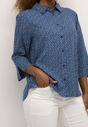 Vrouw draagt een blauw bloemenblouse met knopen en driekwart mouwen en witte broek, met één hand in haar zak.