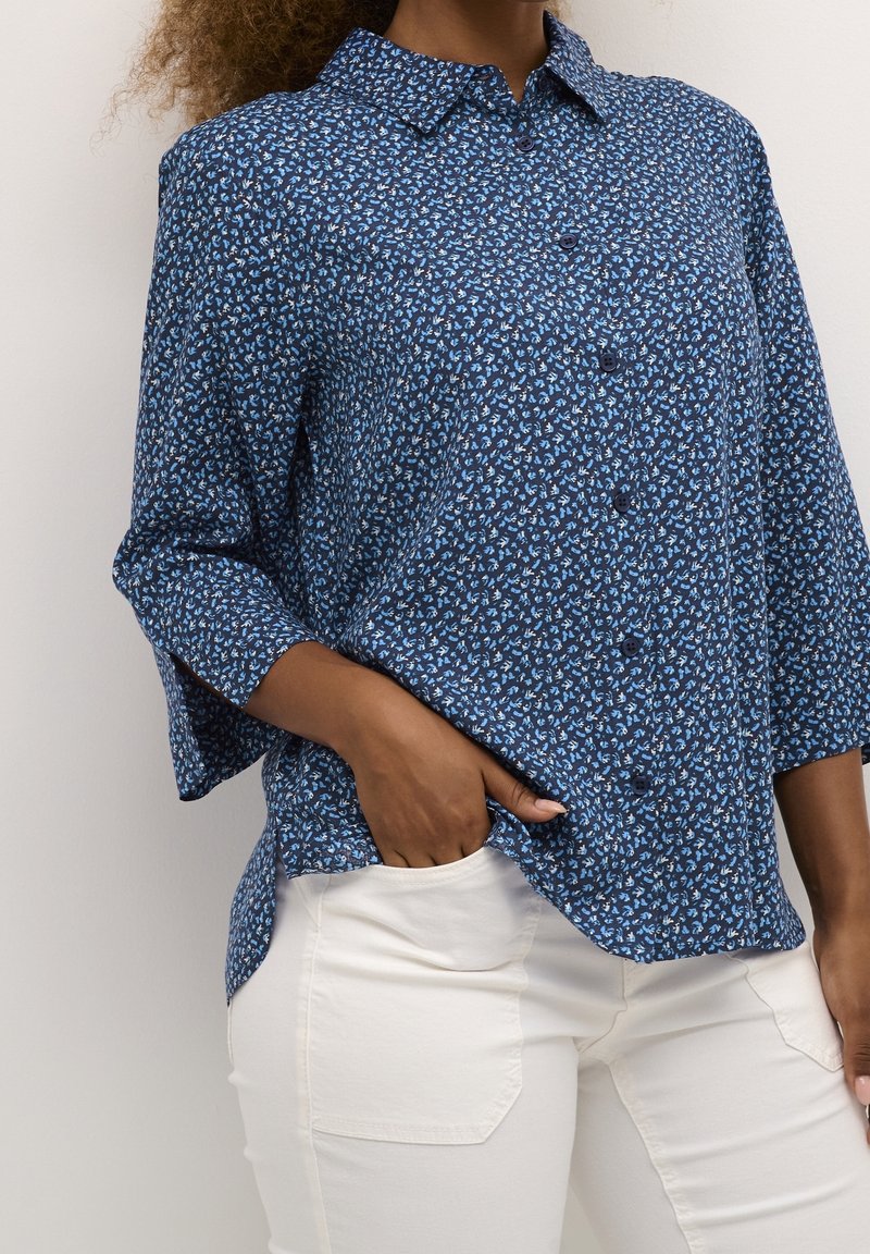 Femme portant une chemise boutonnée bleue à motifs floraux avec manches trois-quarts et un pantalon blanc, une main dans la poche.