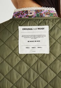 Veste matelassée vert olive avec un col à motifs floraux. Comprend une étiquette de soin carrée avec du texte. Tissu texturé, couture visible.
