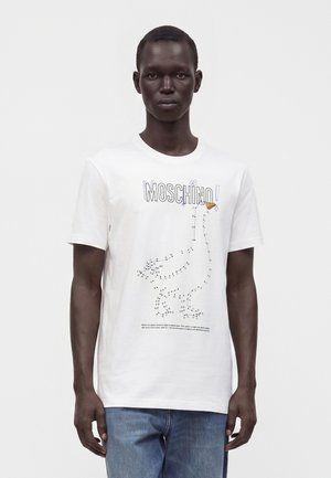 Mand iført hvid Moschino t-shirt med et prik-til-prik-motiv af en and og blå jeans mod en ensfarvet baggrund.