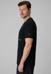Homme aux cheveux courts et bruns avec une légère barbe, portant un T-shirt noir à manches courtes, debout de profil devant un fond gris uni.