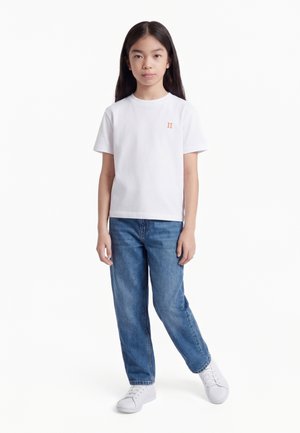 Hosszú hajú gyermek, fehér pólót visel, amelyen apró narancssárga logó található, kék farmert és fehér tornacipőt visel, fehér háttér előtt áll.