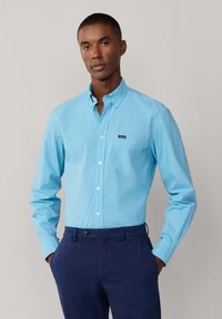 Camisa azul claro abotonada con un bolsillo y un pequeño logo en el pecho, combinada con pantalones azul marino, mostrando una textura de tela lisa.