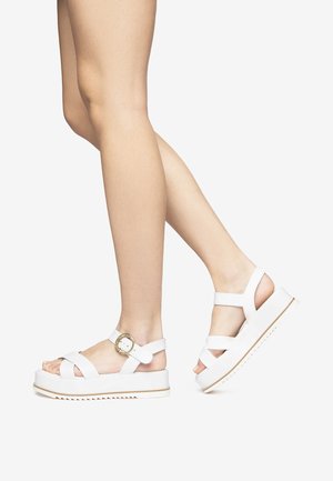White platform sandals with cross straps, a gold buckle accent, and a textured sole. Smooth material, modern design, and open heel.