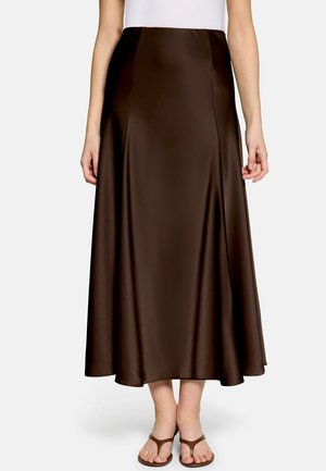 Femme portant une jupe midi en soie brun foncé taille haute, associée à des sandales marron à lanières, debout sur un fond uni.