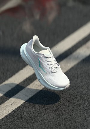 White athletic shoe with textured upper and turquoise accents. Rounded sole, lace-up design, displayed mid-air over a dark surface.